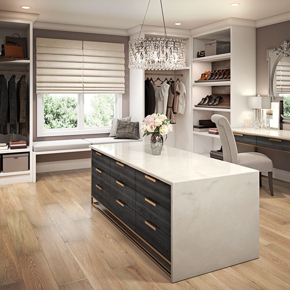 Large walk-in closet with island of white quartz top & sides and dark wood drawers, wood look tile flooring, window seat, makeup vanity, clothing racks, shoe and accessory shelves.