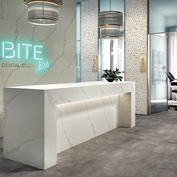 Dentist office reception desk with white & gray quartz slab wall and waterfall counter, teal neon sign, gray stone flooring tile, and white & brass mosaic accent wall.