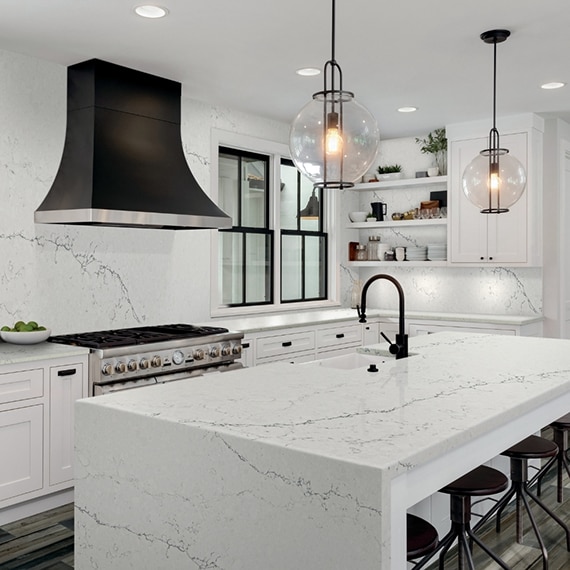 Modern kitchen with white & gray veining stone-look countertops, waterfall island, and matching slab backsplash, black vent hood, and globe pendants.
