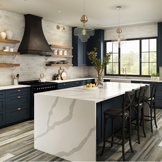 Modern farmhouse kitchen with marble look quartz countertops and waterfall island, cream subway tile, navy cabinets, wooden floating shelves, aged wood look tile flooring.