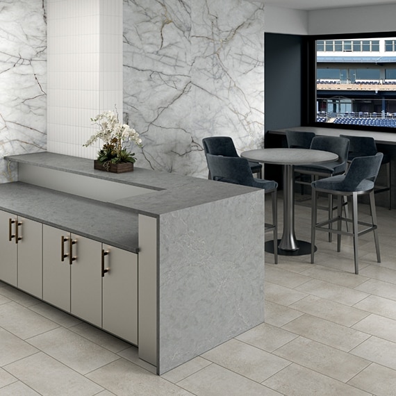 Stadium suite with beige stone look floor tile, gray quartz waterfall countertop, walls covered with porcelain slab that looks like white marble with gray & gold veining.