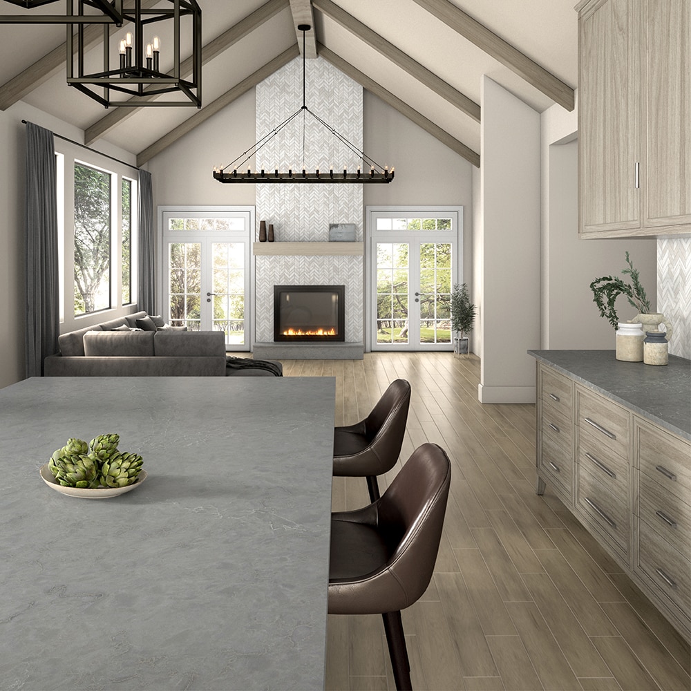 Kitchen with dining room table, living room in background, gray stone look countertops, light wood cabinets, light wood floors, and white walls.