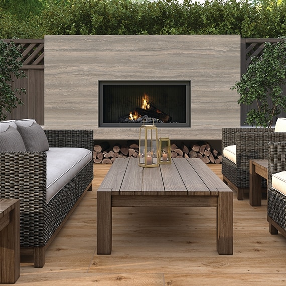Outdoor patio fireplace with beige porcelain slab surround that looks like travertine, wicker sofa & chair with gray cushions, and wooden coffee table.