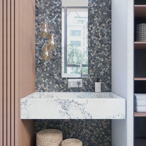 Bathroom vanity with gray pebble mosaic backsplash, mirror, polished silver wall-mounted faucet, white & gray quartz sink, and glass pendant lighting.