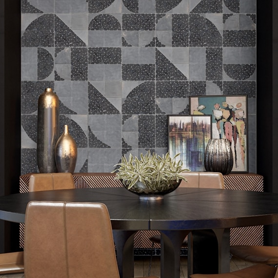 Dining room with gray and black geometric wall tile, buffet holding gold vases & small paintings, round wooden table with brown leather chairs.