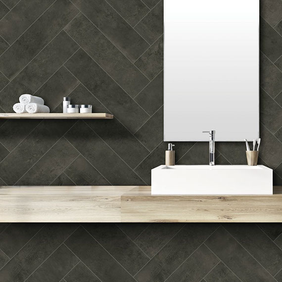 Bathroom backsplash of charcoal gray wall tile in a herringbone pattern with mirror and floating shelf, white vessel sink on floating natural wood vanity.