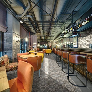 Restaurant with gray & beige encaustic floor tile, wood tables with bright mismatched chairs, encaustic wall tile, brick columns, and ceiling with exposed pipes & ductwork.
