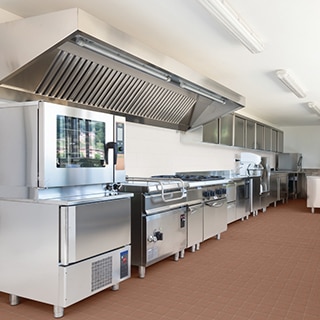 Restaurant kitchen with red quarry floor tile, stainless steel oven, stove, hood vent, ice maker, upper cabinets, and florescent lamps.