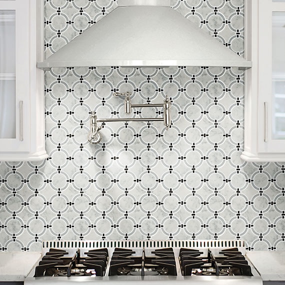 Kitchen with backsplash of white natural stone marble linked ring mosaic tile, gas stove top with white vent hood, silver pot filler, and white cabinets.