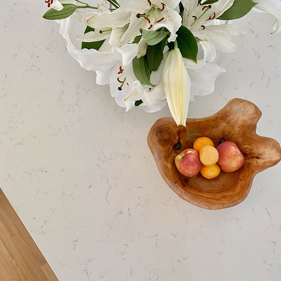 Closeup of white quartz countertop with gray veining, natural wooden bowl holding apples & oranges, and bouquet of white lilies.