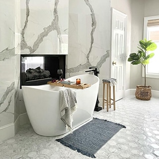 Bathroom with freestanding bathtub, wall-mounted fireplace, white marble look porcelain wall slab with gray veining, and white hexagon marble floor tile.