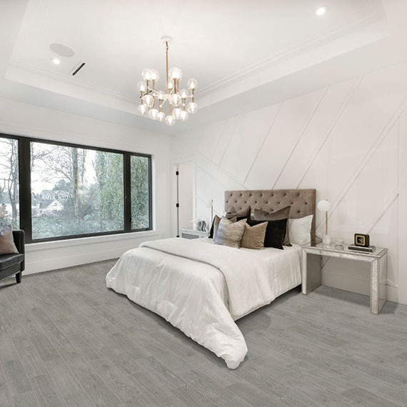 Bedroom with gray floor tile that looks like wood, large picture window, bed with tan tufted headboard, beige pillows, white comforter, and mirrored furniture.