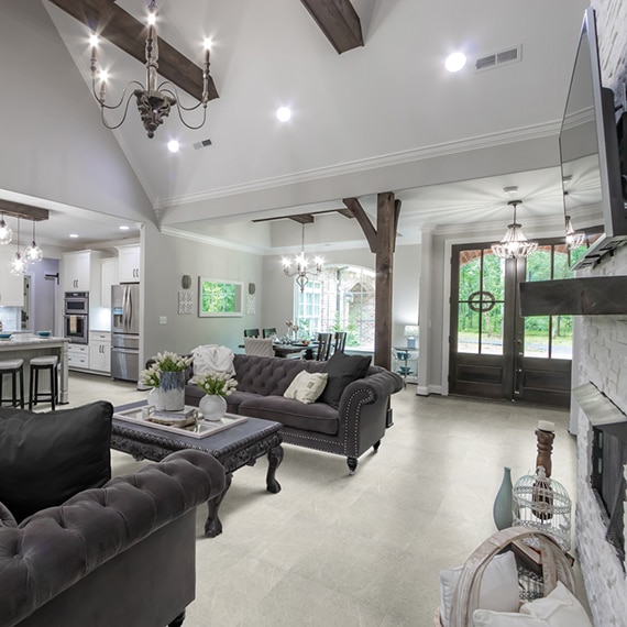 Modern farmhouse open kitchen, living and dining room with off-white marble-look tile, wood beams, pillars, and mantel, and gray velvet couches.