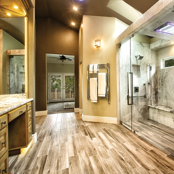 Spacious bathroom with walk-in glass shower of off-white & brown porcelain slab that looks like marble, long mirrored vanity with granite counter, and wood-look floor tile.