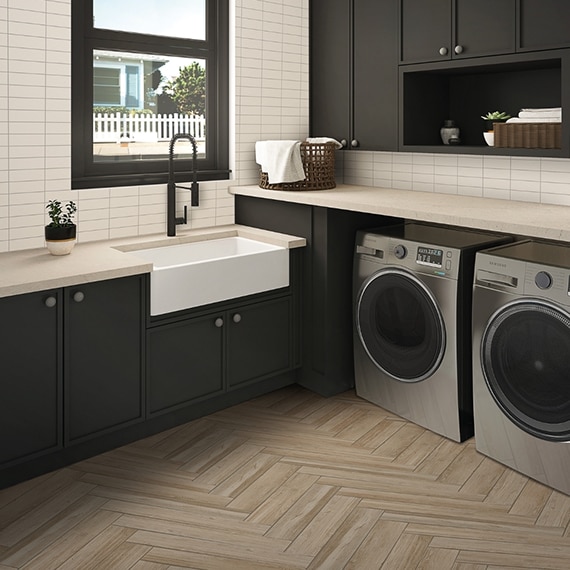 Laundry room with wood look floor tile in herringbone pattern, front-loading washer and dryer, brown cabinets, off-white quartz counters, and white subway wall tile.