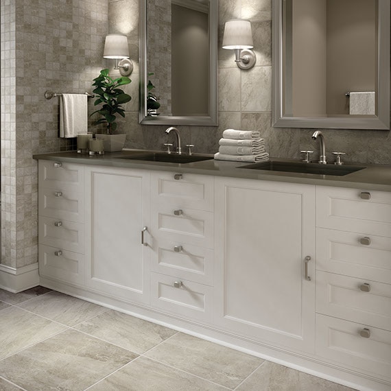 Bathroom double vanity with white cabinetry, framed mirrors, nickel hardware, and white shaded sconces. Warm grey stone-look twelve by twenty four tile on the floor.
