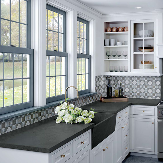 Kitchen with black soapstone countertops, black farm sink in front of windows, geometric backsplash, and white cabinets.
