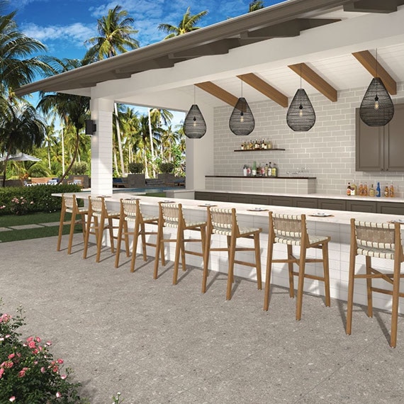 Resort outdoor bar with off-white subway tile backsplash, beige slab countertop, white tile base, wooden bar stools on brown pavers that look like stone.