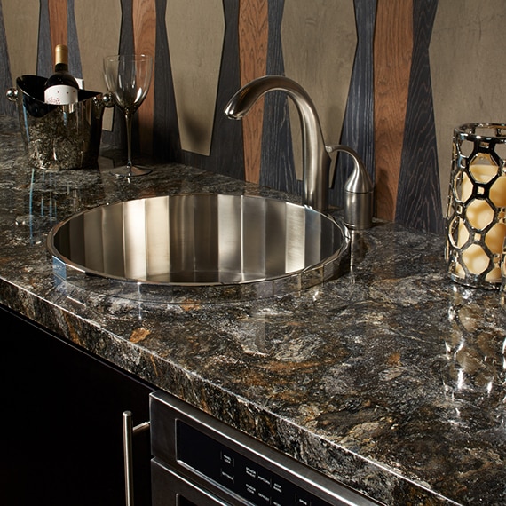 Wet bar with dark granite gray and rust countertop, stainless steel faucet and sink, tan backsplash with black & wood grain accents.