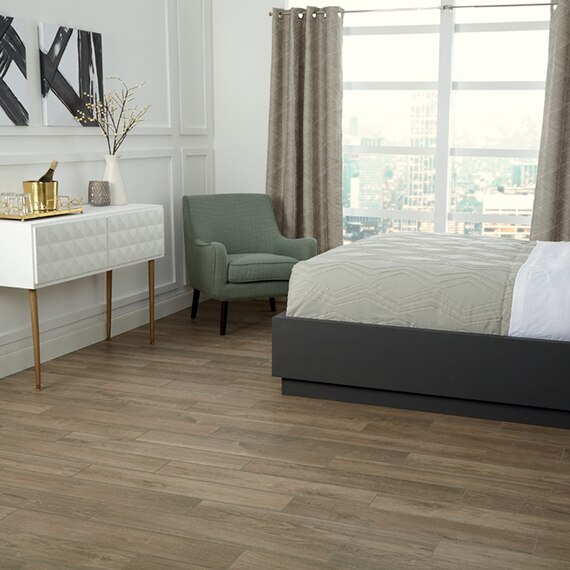 Hotel room with floor tiles that look like wood flooring, platform bed with olive comforter, green chair, white side table, and ice bucket with champagne.