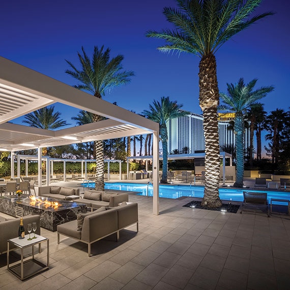 Condominium pool with blue tile, deck of large format taupe tile that looks like stone, palm trees, covered patio with firepit surrounded by tan couches.