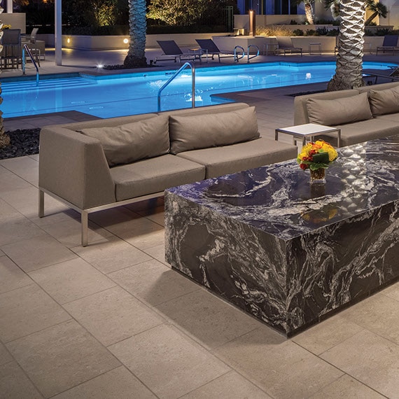 Condominium pool with blue tile, deck of large format taupe tile that looks like stone, palm trees, covered patio with tan couches.