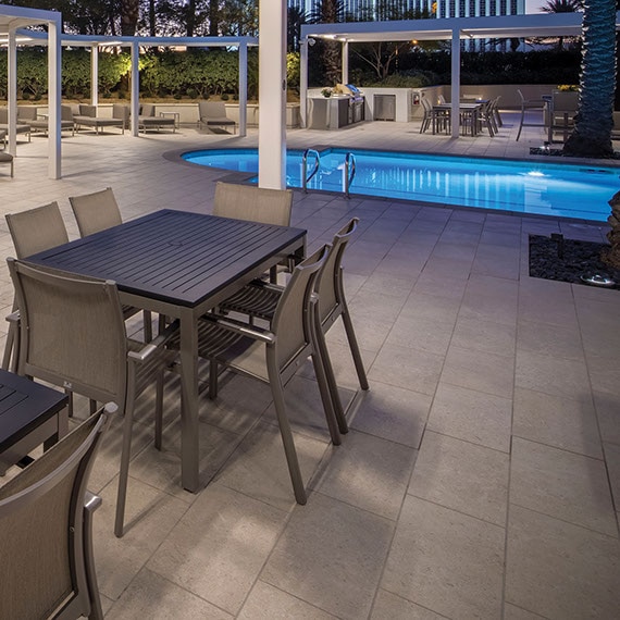 Condominium pool with blue tile, deck of large format taupe tile that looks like stone, palm trees, covered patio with table and chairs.