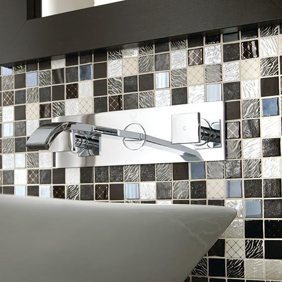 Closeup of off-white, gray, and brown glass mosaic backsplash and polished silver wall mounted faucet over white vessel sink. 