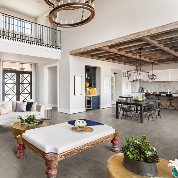 Modern farmhouse living room with stone look tile floor and white furniture, and open kitchen with black table & chairs, and wood beams across the ceiling.