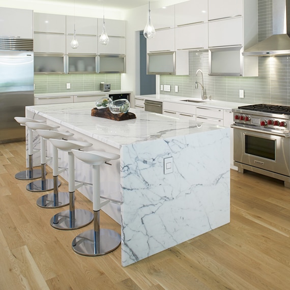 Modern kitchen with white & gray waterfall island, white & silver bar stools, wood look floor tiling, white quartz countertops, light green glass backsplash, and white cabinets.