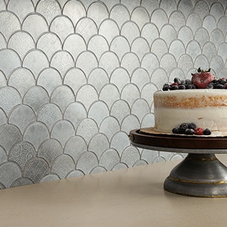 Close up of kitchen backsplash with silver fish scale mosaic tile and smooth beige countertop. Cake on stand with berry decorations sitting on top.