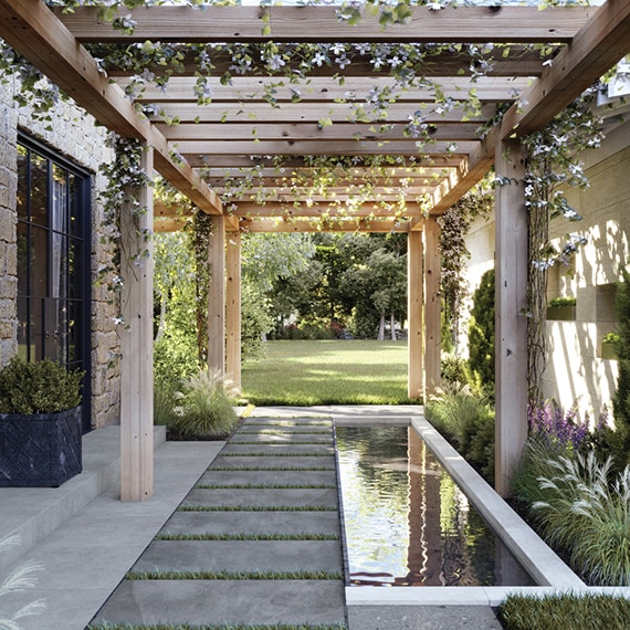 Outdoor courtyard with gray 2CM stone look porcelain pavers, coy pond surrounded by fountain grass, covered by a natural wood pergola with flowering vines.