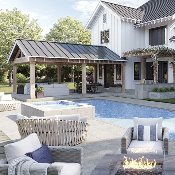 Modern farmhouse two-story home with white wood siding, black metal roof, pool with blue tile & gray deck that looks like stone, covered patio with couch and dining table.