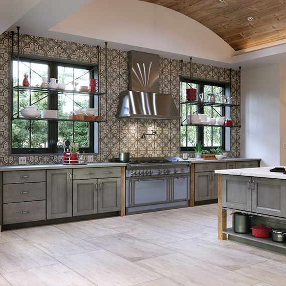 Country style kitchen with gray & beige encaustic wall tile, floating shelves over picture windows, floor tile that looks like wood, and gray porcelain slab countertops.