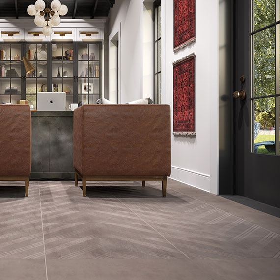 Home office with desk and leather chairs in the background, large windows on the right, and textured taupe porcelain tile in a chevron pattern on the floor.