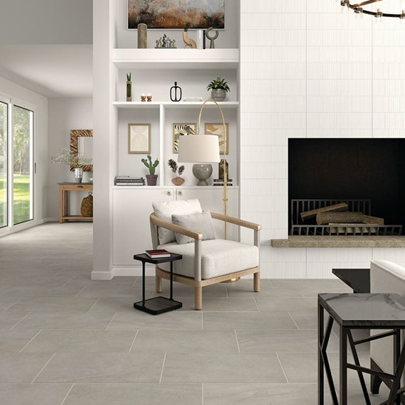 Living room with gray floor tile that looks like stone, fireplace with white textured tile surround, wood chair with off-white cushions.