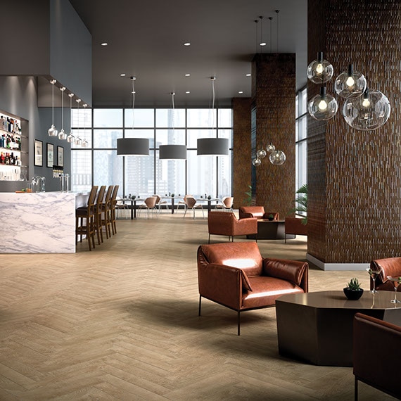 Spacious hotel lobby and bar with wood-look tile floor in a herringbone pattern. Light brown leather modern style low arm chairs and coffee tables on the side.
