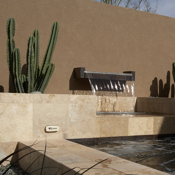 Rustic outdoor pool, spa, and fountain with tan travertine natural stone, aloe, saguaro and senita cactus in ground covered with rocks.