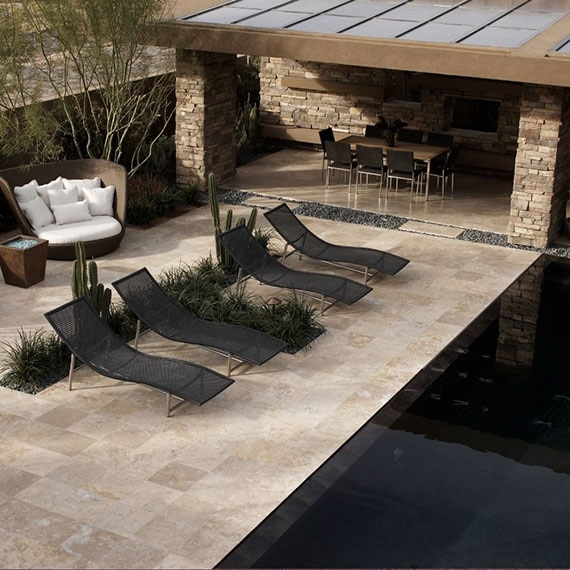 Rustic pool with tan travertine natural stone deck, black lounge chairs, wicker loveseats next to firepit, table & chairs under covered patio.