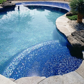 Tropical pool with iridescent blue tile, light gray paver deck, lava rocks, and palm trees set in gravel.