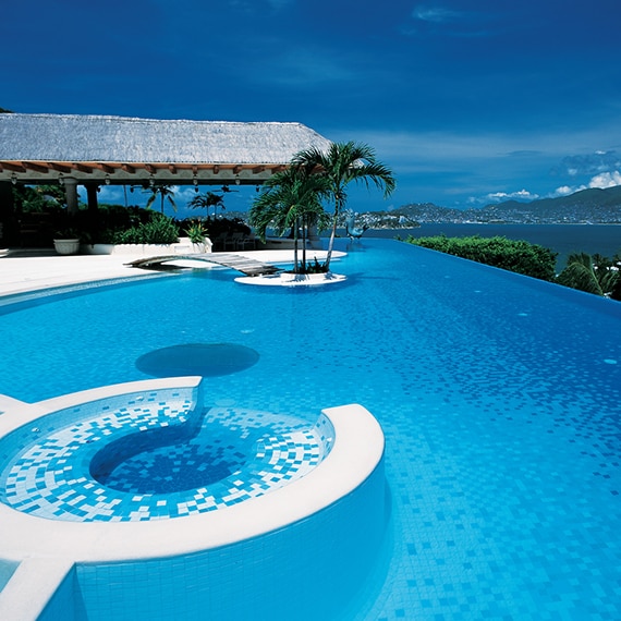 Infinity pool with white and blue tile, wood bridge to swim-up island with palm trees, patio with thatched roof, ocean view with mountains in the background.