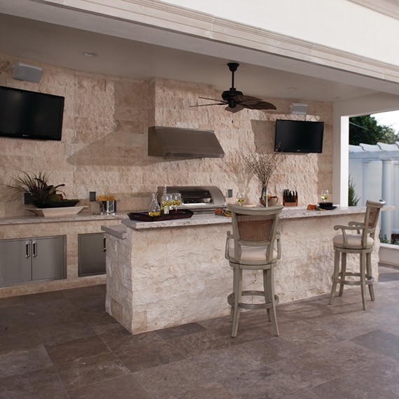 Rustic outdoor kitchen with coral travertine natural stone wall & island base, wood & wicker bar stools, brown travertine floor tile, and stainless steel appliances.