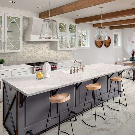 Modern farmhouse kitchen with wood beams, taupe stone-look mosaic backsplash, tan quartzite countertops & island, and gray marble look floor tile
