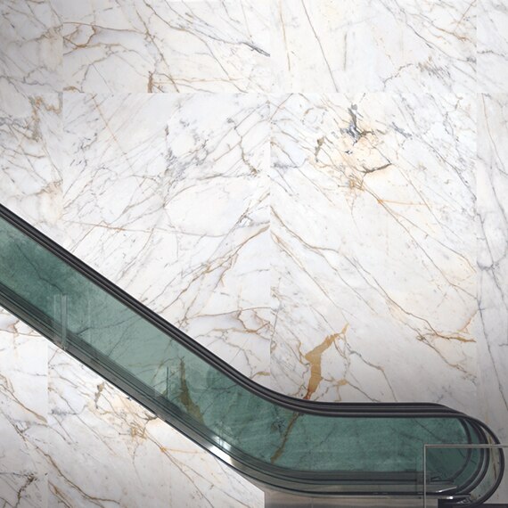 Escalator with green-tinted glass railing, large-format porcelain slab wall that looks like white marble with gold and gray veining.