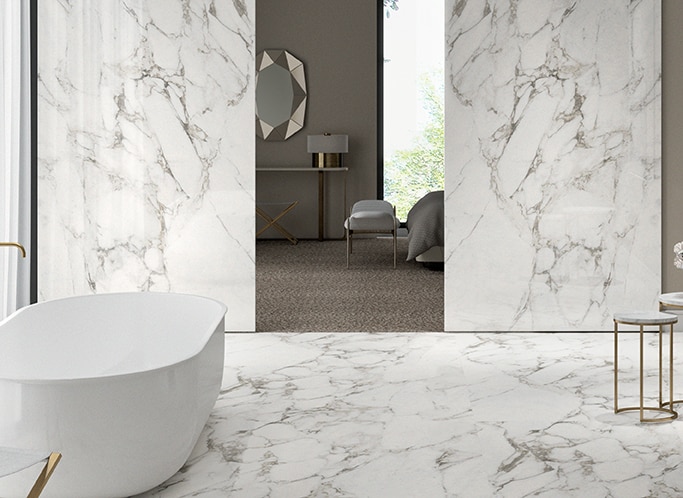 Bathroom with white bathtub, white marble look slab on floor and walls, and bedroom seen through the doorway.