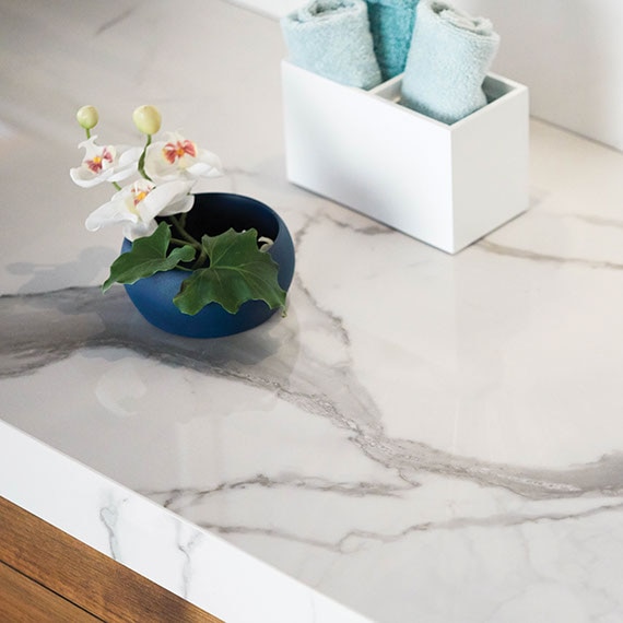 Closeup of bathroom vanity with white marble-look porcelain slab countertop on natural wood cabinet and white hibiscus in blue pot.