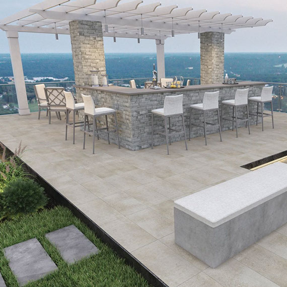 Rooftop patio with stacked stone-faced bar under a pergola, wood look slip-resistant floor tile, and gray paver walkway set in grass.