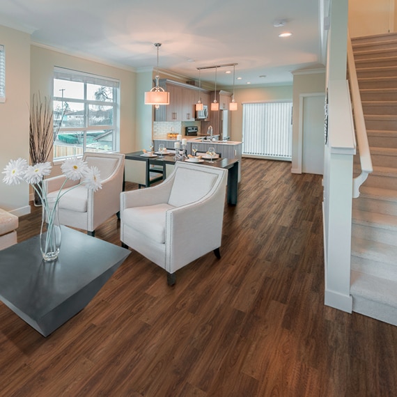 Open concept living room, dining room, and kitchen with luxury vinyl tile that looks like wood flooring, white cushioned chairs, black table & chairs, pendants over kitchen peninsula.