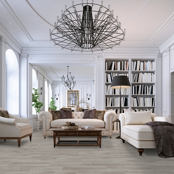 Home library with light wood look tile flooring, white overstuffed sofa & chairs, floor-to-ceiling bookshelves, and metal chandeliers.