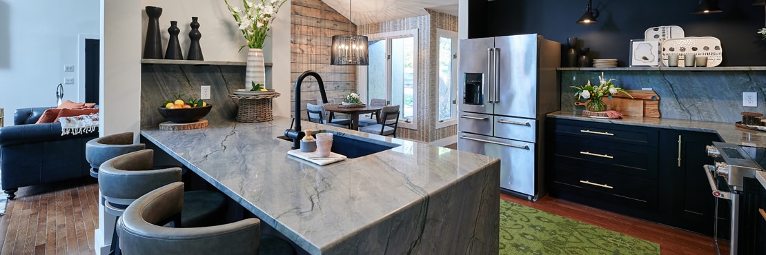 Kitchen with gray natural quartzite island with sink and waterfall countertop, backsplash and floating shelves, and lower black cabinets.